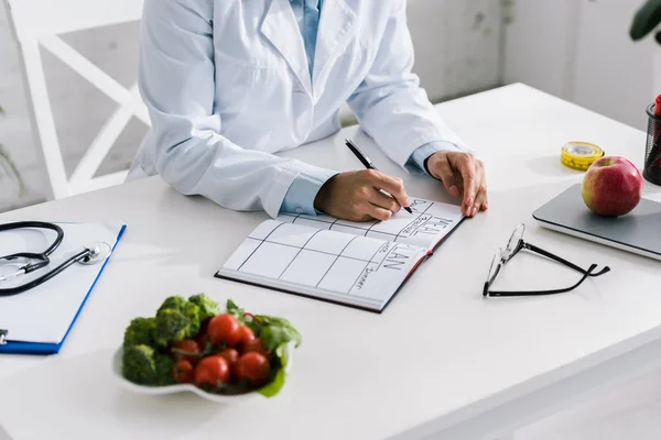 Vue recadrée de l'écriture nutritionniste dans un cahier près des légumes et des pommes — Photo de stock