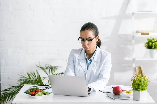 Attraktive Ernährungsberaterin in Brille mit Laptop in der Nähe von Gemüse und Pflanzen — Stockfoto