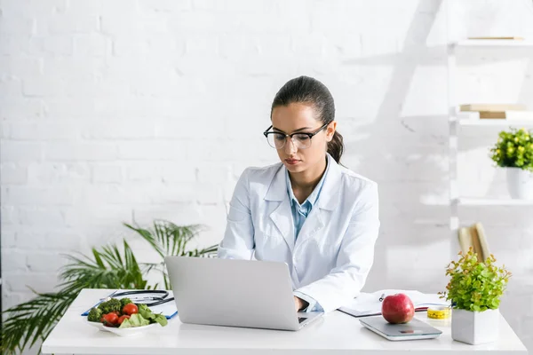 Nutricionista atractiva en gafas usando portátil cerca de verduras y plantas - foto de stock