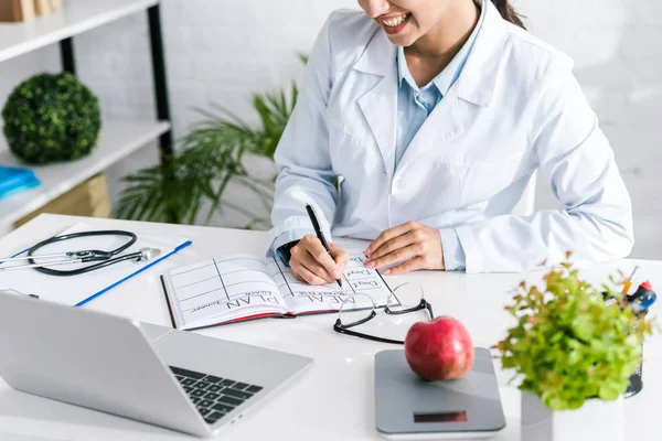 Cropped view of happy doctor writing meal plan in notebook — Stock Photo