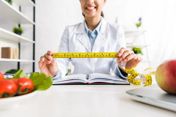 Vista recortada de mujer alegre sosteniendo cinta métrica cerca de verduras y manzana - foto de stock