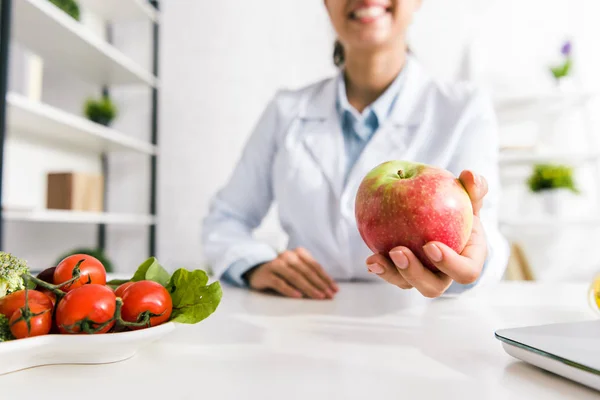 Cropped view of nutritionist holding tasty apple near vegetables — Stock Photo