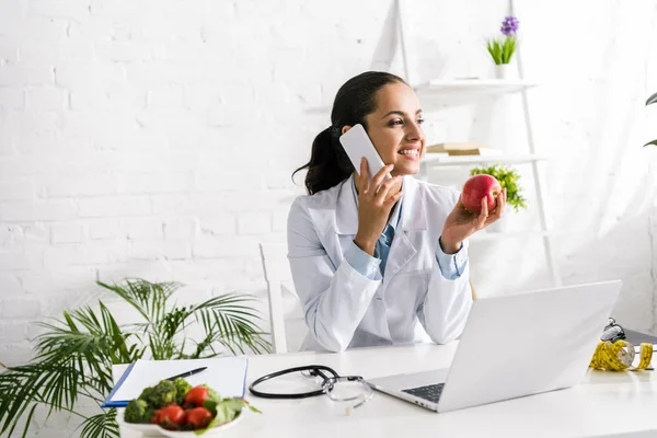 Nutricionista feliz hablando en el teléfono inteligente cerca de la computadora portátil y manzana - foto de stock