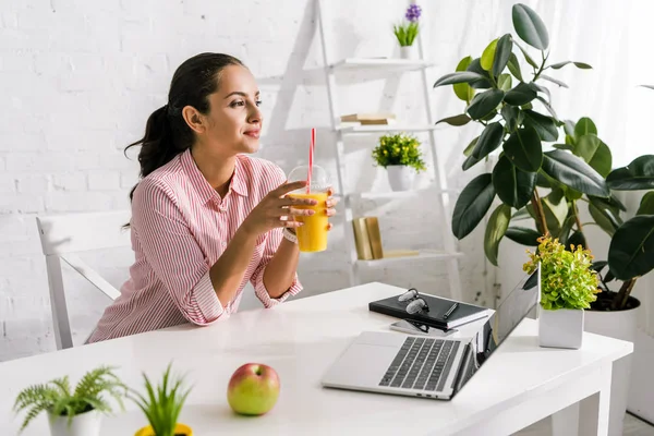 Mujer alegre sosteniendo taza de plástico con jugo de naranja cerca de la computadora portátil y manzana - foto de stock