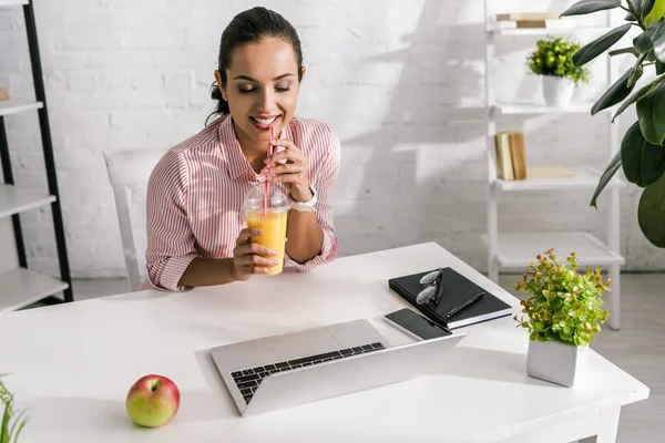 Счастливая женщина держит пластиковую чашку с апельсиновым соком рядом ноутбук и яблоко — стоковое фото