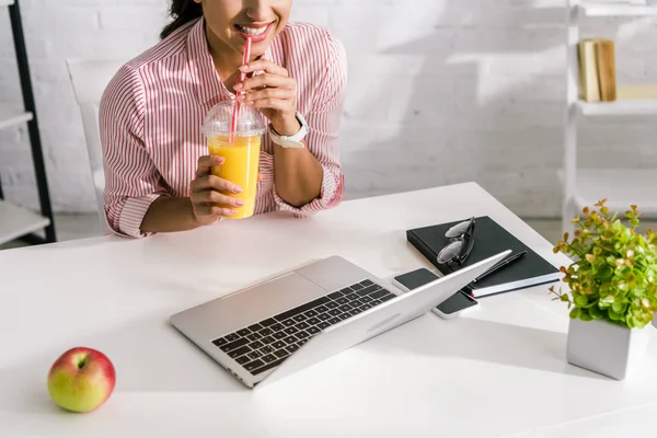 Vista recortada de niña sosteniendo jugo de naranja cerca de la computadora portátil - foto de stock