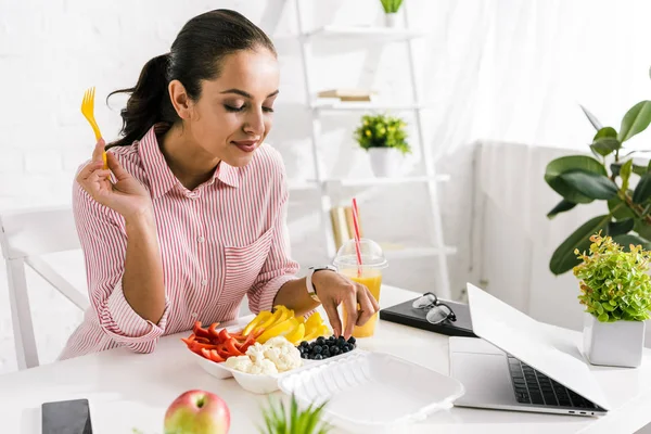 Cheerful woman holding plastic fork near vegetables and gadgets — Stock Photo