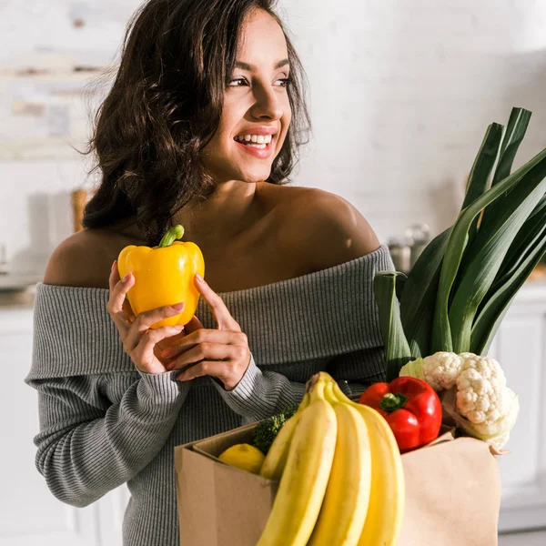 Fille souriante tenant paprika jaune près de bananes dans un sac en papier — Photo de stock