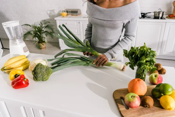 Vista recortada de niña sosteniendo puerro cerca de verduras y frutas en la mesa - foto de stock