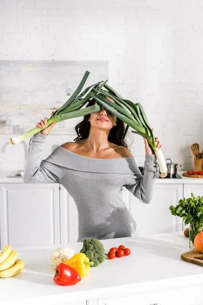 Chica atractiva sosteniendo puerro cerca de la cara y verduras sabrosas en la mesa - foto de stock