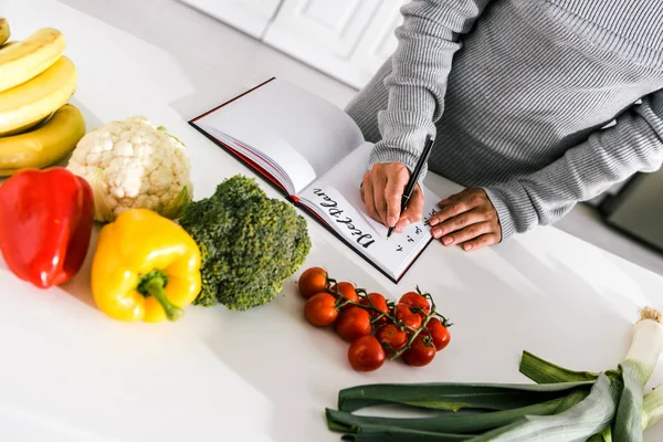 Обрезанный вид женщины, которая пишет в блокноте рядом с овощами — стоковое фото