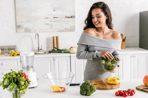 Fröhliche junge Frau hält Grün in der Hand und blickt auf Schüssel mit Salat — Stockfoto