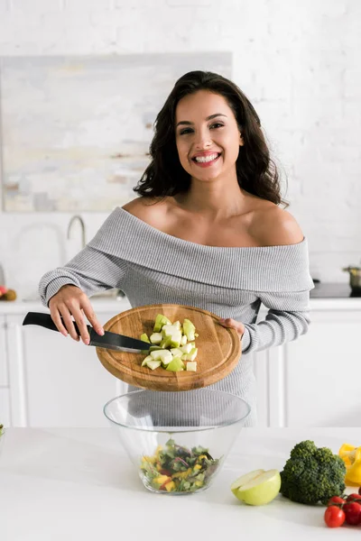 Happy girl holding knife and cutting board near salad in bowl — Stock Photo