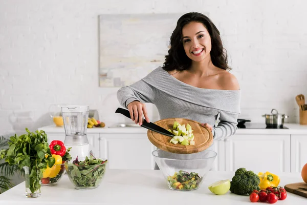 Fröhliches Mädchen hält Messer und Schneidebrett neben Salat in Schüssel — Stockfoto