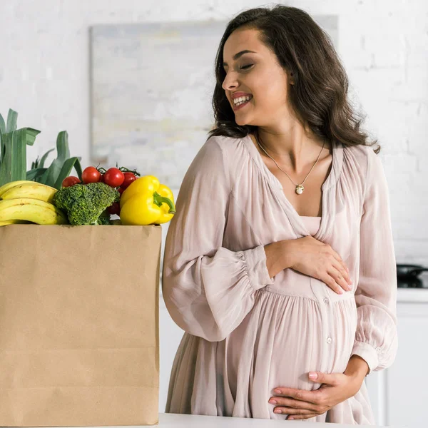 Весела вагітна жінка дивиться на паперовий мішок з продуктами, торкаючись живота — стокове фото