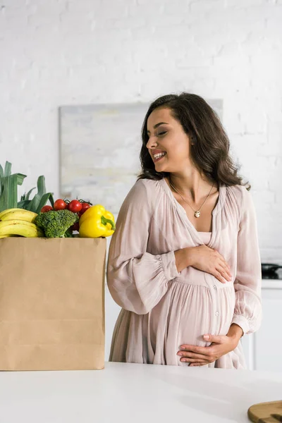 Щаслива вагітна жінка дивиться на паперовий мішок з продуктами, торкаючись живота — стокове фото