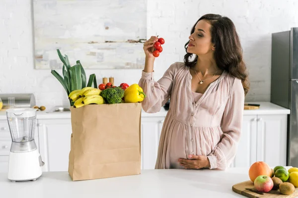 Mujer embarazada feliz mirando tomates cherry cerca de bolsa de papel - foto de stock