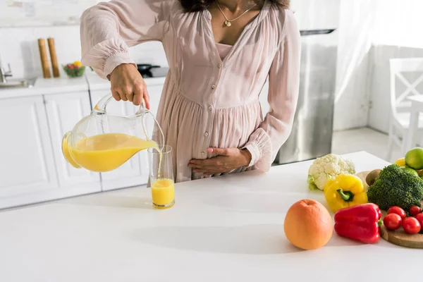 Vista ritagliata della donna incinta versando succo d'arancia in vetro vicino alle verdure — Foto stock