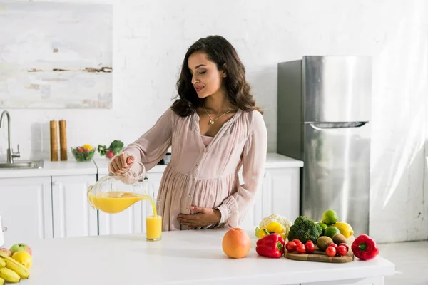 Hermosa mujer vertiendo jugo de naranja en vidrio cerca de verduras - foto de stock