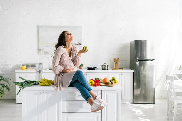 Happy pregnant woman holding apple while sitting on table — Stock Photo