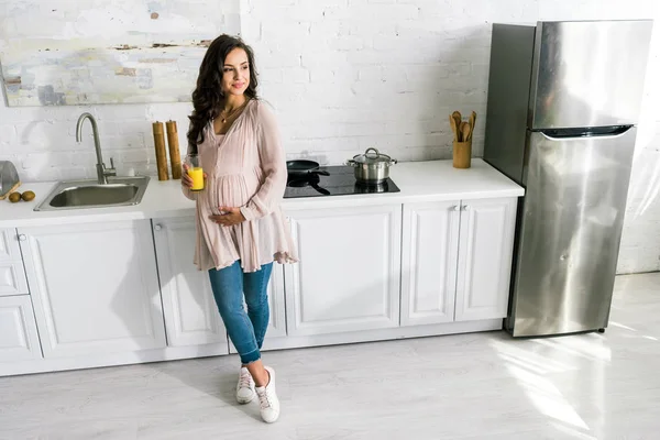 Fröhliche schwangere Frau steht und hält Glas mit frischem Orangensaft in der Küche — Stockfoto