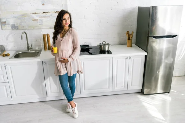 Cheerful pregnant woman standing and holding glass with fresh orange juice in kitchen — Stock Photo