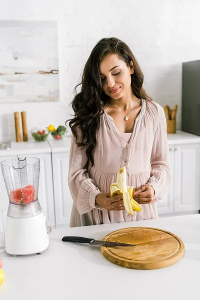 Mulher grávida descascando banana perto do liquidificador com toranja — Fotografia de Stock