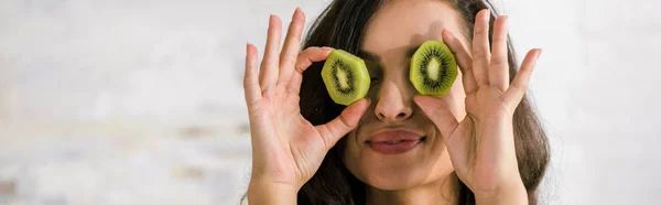 Panoramic shot of happy woman holding halves of kiwi fruit while covering eyes — Stock Photo