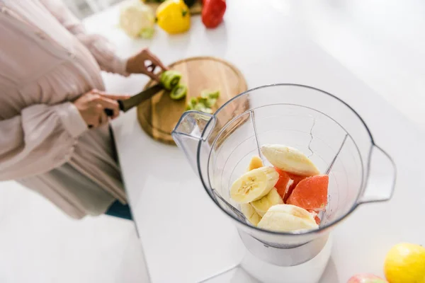 Foyer sélectif du mélangeur avec des fruits près de la femme enceinte coupe kiwi — Photo de stock