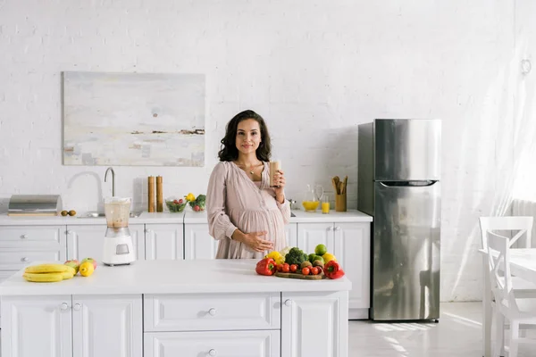Привлекательная беременная женщина держит стакан со смузи возле стола — стоковое фото