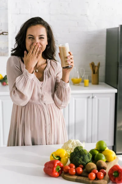 Gaie femme enceinte tenant verre avec smoothie savoureux et couvrant visage — Photo de stock