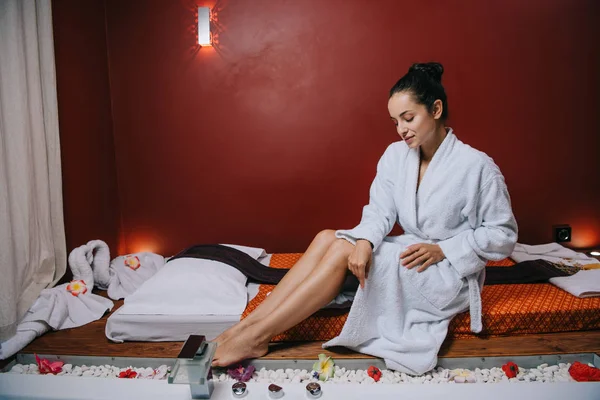 Atractiva mujer en albornoz sentado en alfombra de masaje en el salón de spa - foto de stock