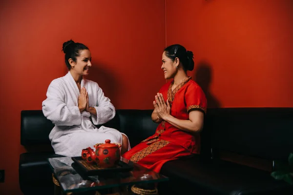 Улыбающаяся женщина и азиатский массажист сидят на диване и показывают приветственные руки — стоковое фото