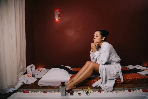Atractiva mujer con los ojos cerrados en albornoz sentado en la alfombra de masaje en el salón de spa - foto de stock