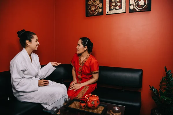 Lächelnde Frau und asiatischer Masseur sitzen auf dem Sofa und unterhalten sich im Wellnessbereich — Stockfoto