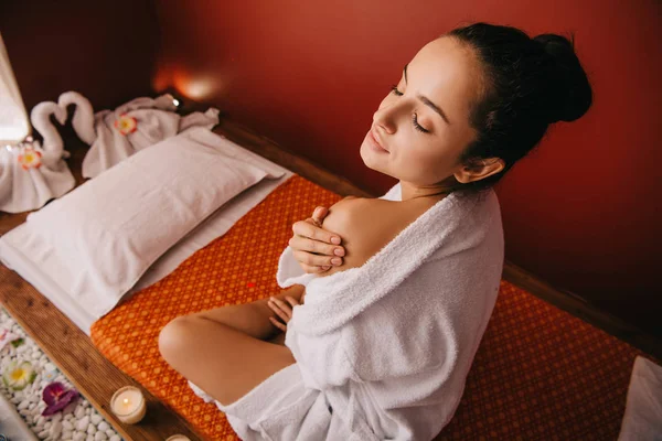 Vista de ángulo alto de la atractiva mujer en albornoz sentado en la alfombra de masaje en el salón de spa - foto de stock