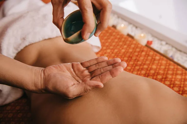 Vista cortada do massagista derramando óleo de fragrância na mão e mulher deitada no tapete de massagem — Fotografia de Stock