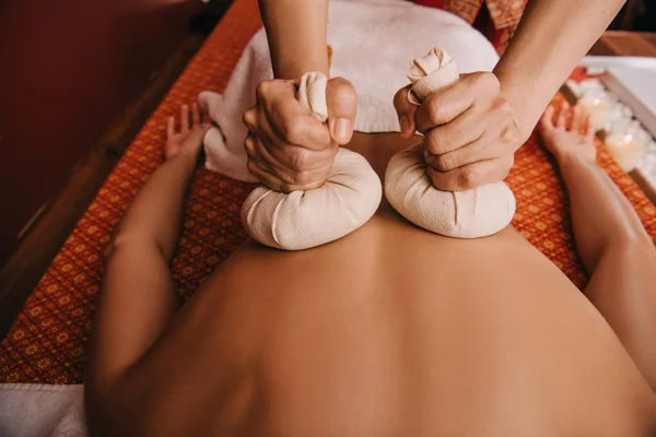 Vista cortada de massagista fazendo massagem nas costas com bolas de ervas para a mulher no spa — Fotografia de Stock