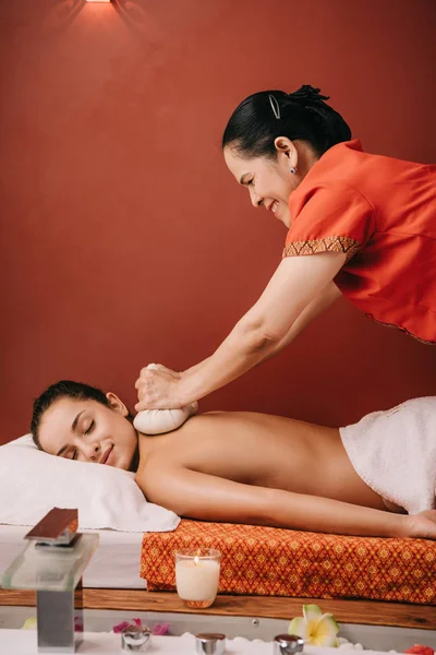 Vista lateral de masajista asiático haciendo masaje de espalda con bolas de hierbas a mujer en spa - foto de stock