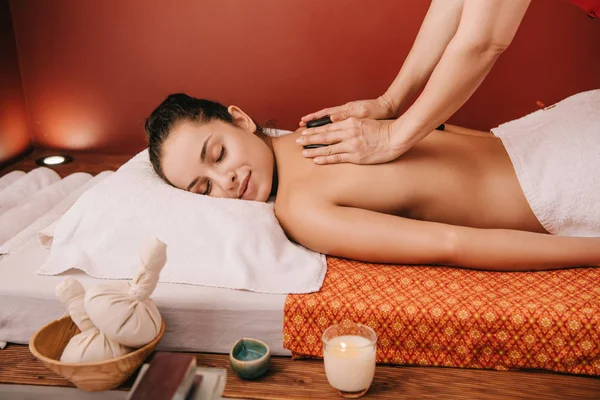 Vista recortada de masajista haciendo masaje de piedra caliente a la mujer en el salón de spa - foto de stock