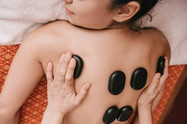 Vista ritagliata del massaggiatore facendo massaggio con pietre calde alla donna nel salone spa — Foto stock