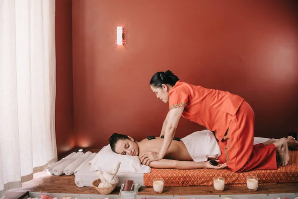 Vue latérale de masseur asiatique faisant massage aux pierres chaudes pour femme dans le salon de spa — Photo de stock