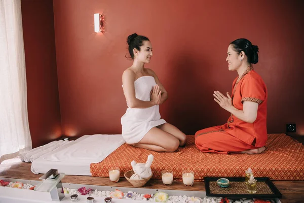 Sonriente mujer y asiático masajista sentado en masaje mat y mostrando saludos manos - foto de stock