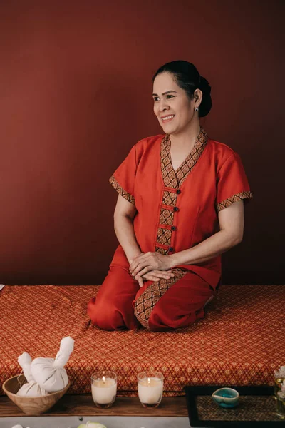 Азиатский массажист в красном костюме улыбается и сидит на коврике для массажа — стоковое фото