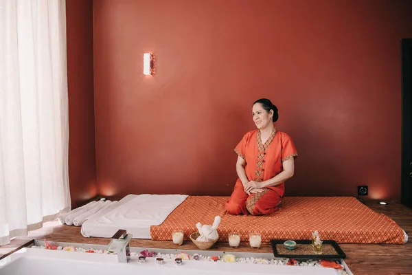 Азіатський масажист в червоному костюмі посміхається і сидить на масажному килимку — стокове фото