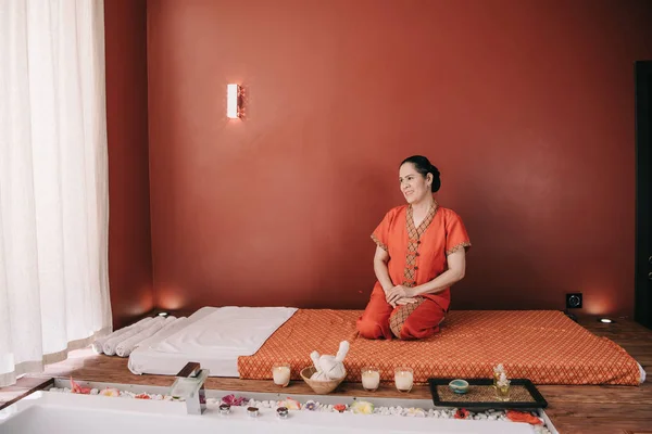 Asian masseur in red costume smiling and sitting on massage mat — Stock Photo