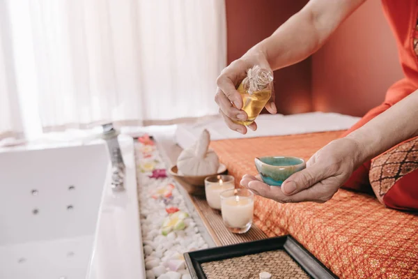 Vista cortada do massagista sentado no tapete de massagem e derramando óleo de fragrância para tigela — Fotografia de Stock