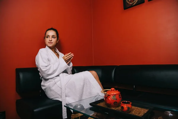 Привлекательная женщина в белом халате сидит на диване и держит чашку — стоковое фото