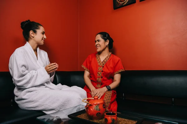 Smiling woman in bathrobe and asian masseur talking in spa — Stock Photo