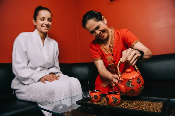 Азіатський масажист наливає чай на чашку і приваблива жінка в халаті посміхається в спа-центрі — стокове фото